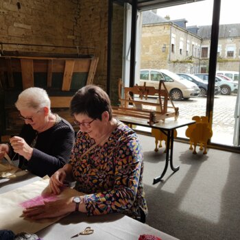 Deux femmes sont assises côte à côte, concentrées sur des travaux de couture et de broderie autour d'une table parsemée de pelotes de laine colorées.