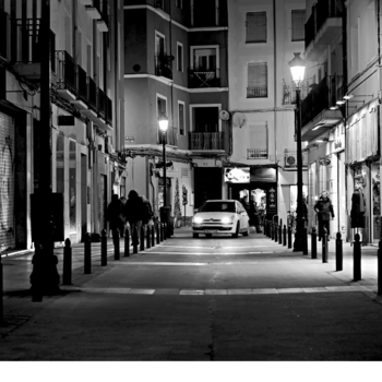 Une vue nocturne en noir et blanc d'une rue étroite, bordée de bâtiments et délimitée par des bornes, avec une voiture circulant au loin et quelques passants.