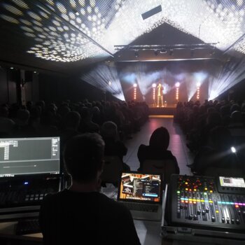 Une vue depuis la régie montrant un technicien manipulant une console de mixage et des écrans lors d'un spectacle en salle, avec un artiste sur scène sous des projecteurs.