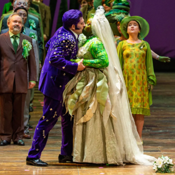 Sur une scène de théâtre, un homme en costume violet embrasse une femme vêtue d'une robe de mariée aux teintes vertes.