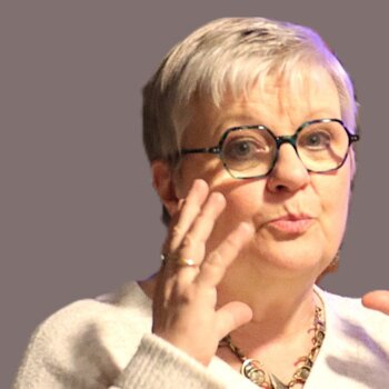 Portrait d'une femme aux cheveux courts portant des lunettes et un pull blanc captée dans un geste expressif de la main alors qu'elle semble parler.