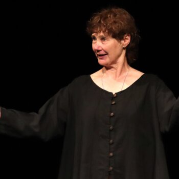 Une femme aux cheveux courts vêtue d'une longue tunique noire se tient sur une scène au fond noir, les bras largement ouverts dans un geste expressif.