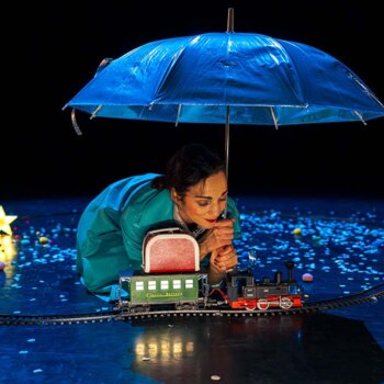 Photo tirée du spectacle montrant une femme vêtue d'un manteau bleu agenouillée sur une scène sombre et s'abritant sous un parapluie bleu. Elle observe un petit train électrique qui circule sur des rails entourés de confettis et d'une étoile lumineuse. 