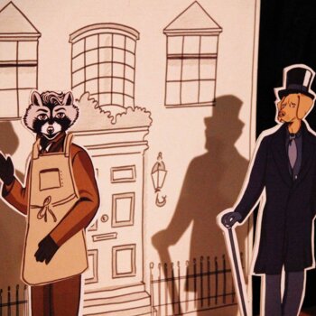 Théâtre de papier avec un raton laveur en tablier et un chien vêtu d'un costume, d'un haut-de-forme et tenant une canne, devant un décor dessiné représentant une façade de maison.