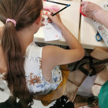 Jeune fille aux longs cheveux bruns attachés en queue-de-cheval assise à une table s'applique à dessiner et écrire sur une carte de vœux où l'on devine le mot « Maman ».