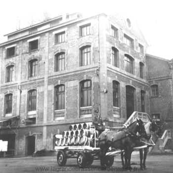 Photographie en noir et blanc d'un bâtiment industriel en briques, devant lequel un attelage de deux chevaux tire une charrette chargée de fûts.