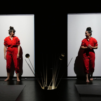 Deux femmes en combinaisons rouges se tiennent debout devant deux grands panneaux blancs verticaux sur une scène sombre, séparées par une structure centrale de tiges métalliques.