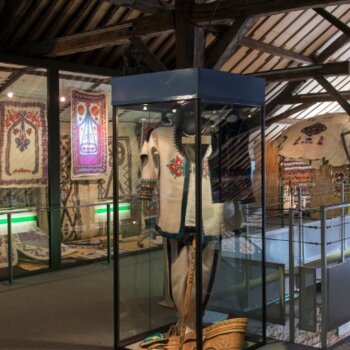 Photographie représentant l'étage du Musée du Feutre situé sous une charpente en bois où sont exposés des costumes et tapis en Feutre blanc brodés derrière de grandes vitrines et dans des présentoirs centraux.