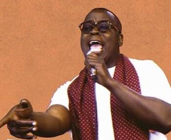 Brother Wallace, un chanteur noir portant des lunettes et une écharpe rouge à pois chante avec passion dans un micro, un bras tendu vers l'avant.