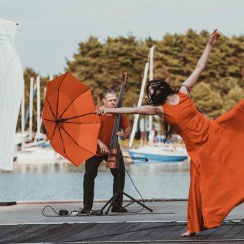 Deux interprètes : une danseuse vêtue d'une robe et d'un parapluie orange et un musicien jouant d'un instrument à corde vêtu d'un t-shirt orange. Décor muni d'un lac sur lequel sont posés des voiliers.