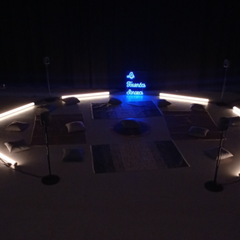 Une installation immersive circulaire au sol, composée de tapis et de coussins, entourée de tubes lumineux et de haut-parleurs, avec une enseigne néon bleue portant l'inscription « Les Tournées Sonores »