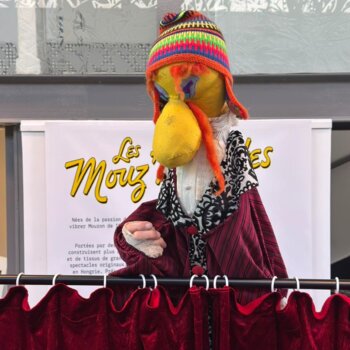 Marionnette colorée à bonnet tricoté, installée derrière un rideau de théâtre rouge.