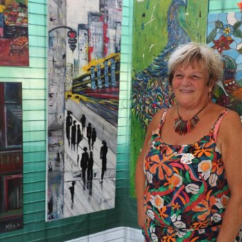 Une femme souriante, portant une tenue fleurie et un collier coloré, pose devant une exposition de plusieurs peintures aux styles variés, incluant des scènes urbaines et des représentations d'animaux.