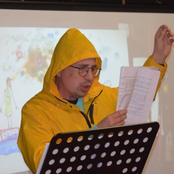 Un homme portant un ciré jaune à capuche et des lunettes lit un texte sur un pupitre, avec une illustration projetée en arrière-plan.